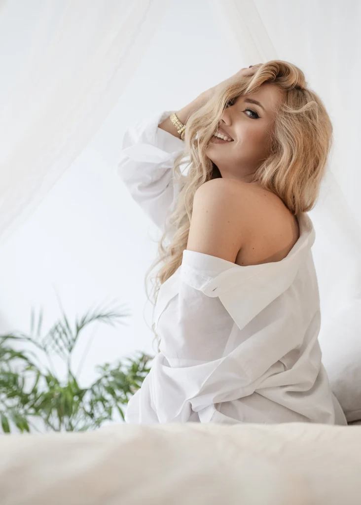 Uśmiechnięta kobieta w białej koszuli na łóżku – sesja sensualna